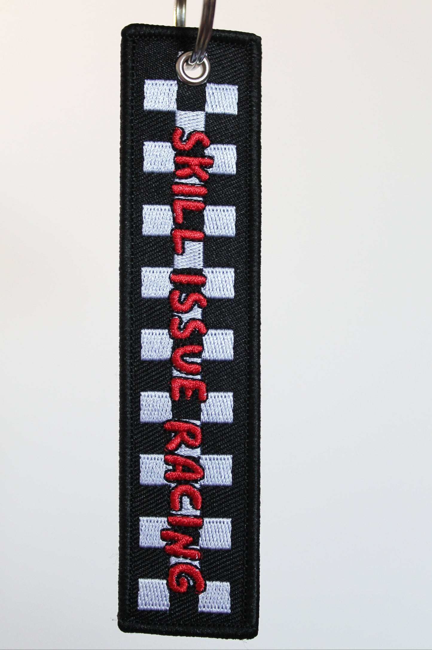 A rectangular embroidered keychain or tag with a black border, white checkered stripes, and red vertical text reading 'SKILL ISSUE RACING' on a white background.